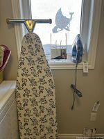 Photo of a folding ironing board with floral pattern cover leaning against a wall near a window. Hamilton Beach Durathon iron mounted next to window.