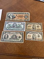 Five banknotes laid out on table: $5 Bank of Commerce, two $1 Bank of Canada, two 25¢ Dominion of Canada.
