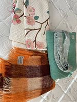 Top down image showing three blankets: cream colored floral quilt, orange and brown striped Glen Cree mohair blanket with fringed ends, and folded sage green Kenwood wool blanket with satin edge.