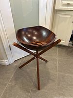 Large dark brown wooden salad bowl with wooden fork and spoon inside, placed on wooden three-legged stand on tiled floor.