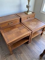 Pair of wooden side tables shown side by side with one table holding a decorative lamp. Tops show parquet wood paneling with visible wear, drawer fronts with metal handles, and open shelves below.