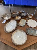 Overview of approximately 12 pieces of silver plated serving ware on wood table including trays, bowls, and cups.