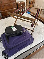 Purple wheeled carry-on luggage with small black travel case and wooden luggage rack on mattress.