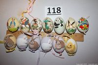 Top view of thirteen hand-painted eggs each with unique animal or character images and attached ribbons, displayed next to ruler for scale