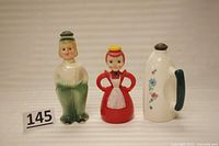Front view of three vintage sprinkle bottles: green and white ceramic male figure, red plastic lady figure, and white ceramic iron-shaped bottle.