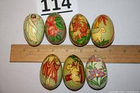 Top view of all seven wooden eggs aligned next to a ruler showing approximate size. Various hand-painted designs including Jesus, lambs, and floral motifs.