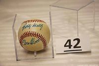 Baseball inside a clear plastic case showing two signatures, displayed with lot number 42 in background.