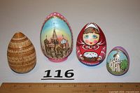 Four wooden Easter eggs displayed side by side: one carved nesting egg in natural wood tone and three painted eggs with Russian imagery including churches and a matryoshka doll.