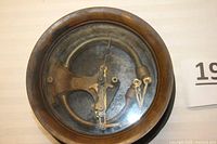 Top view of antique hydraulic gauge with glass cover, showing curved internal tube and mechanical arms.