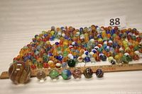Photo shows a large collection of small vintage marbles organized in rows behind a ruler, with a variety of colors and patterns. In front, a row of larger marbles includes a very large marble with red and yellow swirls and other smaller art glass marbles.