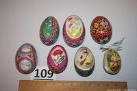 Seven decorated eggs arranged in two rows with a ruler below for size reference, showing diverse motifs and confirming condition and size.