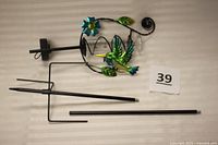 Disassembled pieces of rain gauge laid flat including metal stakes and decorative top portion with hummingbird and flower ornaments.