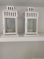 Two white metal and glass lanterns side by side on shelf