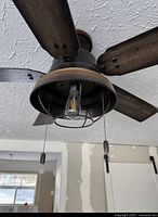 Close-up of fan body showing bronze housing, pull chains, wire cage and bulb, plus wood-look blades