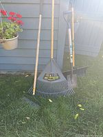 Four yard tools leaning against siding: large plastic leaf rake, shrub rake, snow shovel, spade