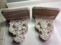 Pair of white carved wooden wall shelves with ornate sconce style floral and scroll carvings placed side by side from an angle showing tops and front design.