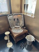 Photo showing two silver trays, two shorter candle holders, a clear glass serving dish, an ice bucket, a small candle snuffer, and two tall champagne flutes with brass stems on and near a small table.