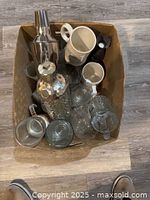Top view of box containing mixed glasses including cocktail shaker, glass bottle, coffee mugs, chalices, and other glassware.