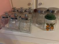 Six hexagonal glass canisters with shiny tin lids, two taller frosted canisters with tin lids, and one smaller square canister with green lid and fruit decoration.
