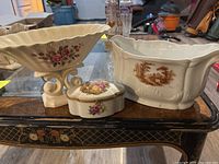 Photo showing three porcelain vessels on a decorative table, including the ceramic pedestal vase with flowers, larger pot-style vase with brown landscape decal, and small ornate jewelry dish with gold accents and floral pattern.