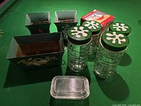 Photo showing three green rectangular tin planters with floral designs, four cylindrical glass storage containers with green lids featuring white flower designs, a clear glass butter dish with lid, and a red box in the background.
