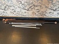 Set of curtain rods including a large black rod with marble finials and five white spring tension rods, displayed on a black surface beneath patterned wallpaper.
