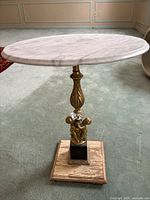 Side view of the marble side table showing round marble top, gold-tone central column, cherub figures, crystal accent, and wooden base with wear.