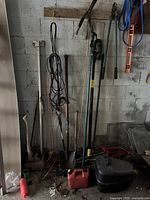 Overall view of garden tools including shovels, axe, and hand tools hanging on a wall and standing on the floor.