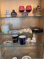 Full shelf display of glassware and porcelain items including cranberry glasses, scent diffuser, mugs, saucers, sugar bowl, and other decorative pieces.