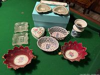 Lot contents including porcelain leaf-shaped dishes, heart-shaped basket dishes, small glass and porcelain dishes displayed on green felt surface.