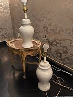 Two cream-colored ceramic table lamps with a grey crackle finish shown side by side on a hardwood floor near floral wallpaper. One lamp is sitting on a wood side table with a gold trim, the other on the floor. Both have bulb fittings; one lamp has a harp for shade attachment.