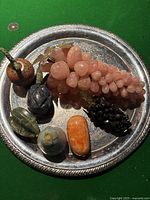 Top view of mixed stone and marble fruit displayed on a silver tray showing various colors and types of carved fruits including grapes and other fruit shapes.