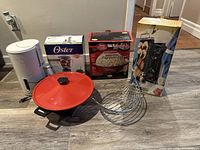 Wide shot showing all items in the lot including white Braun coffee maker, boxed Oster blender, boxed Betty Crocker popcorn popper, boxed cooking stone, electric wok with red lid, and metal fruit basket on floor.