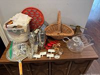Full lot showing mixed kitchen and serving items spread on a wooden cabinet including metal ramekins, glass teapot, wicker basket, plastic container with napkins, red perogie mold, and other small items.