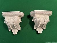 Pair of white ceramic vintage corbels placed side by side on green surface, showing front leaf design patterns.