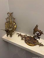 Two brass ornate wall sconce candle holders displayed on corner shelf, showing detailed decorative backs and candle cups