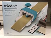 Front of the box showing the Cricut Joy cutting machine cutting gold paper, with smartphone app.