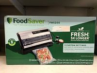 Front of sealed FoodSaver FM5200 box showing vacuum sealing system image, branding, features like 3 custom settings, and included bags and rolls.