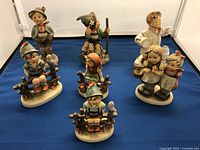 Overview of all seven Goebel porcelain figurines displayed on a blue mat showing detailed poses and colors.