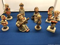 Front view of 8 Goebel Hummel porcelain figurines arranged on blue cloth, showing diversity of characters and poses