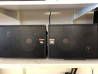 Two black JBL bookshelf speakers with protective metal mesh grills and prominent JBL logos on front. Slight shelf wear visible on bottom shelf.