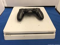 Top view of white Sony PlayStation 4 console with black controller placed on top showing surface wear and logos