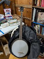 Full banjo with headstock, neck, banjo head, and black carrying case on wooden chair and table with boxes.