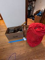 Wide view showing a cardboard box with items inside and a red sleeping bag next to it on hardwood floor