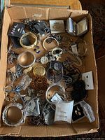 Photo showing a cardboard box filled with assorted costume jewelry including bracelets, pins, earrings, and necklaces in silver and gold tones with some studded stones and beads