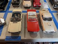 Three vintage model cars displayed on a white surface, showing front and interiors of the cream convertible, red convertible, and cream coupe.