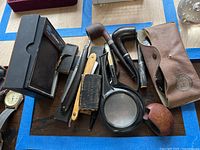 Overall lot on table showing vintage pipes, straight razors, magnifying glasses, and wallet.