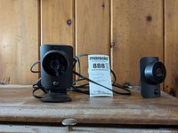 Two black wired security cameras on a wooden surface with a MaxSold lot tag in between