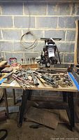Wide shot showing table filled with assorted vintage hand tools including wrenches, clamps, files, and hatchet, with a small anvil on the table