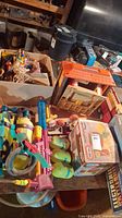 Wide view showing boxed and loose vintage toys including large yellow and purple Playskool water blaster backpack, dolls, and toy boxes.